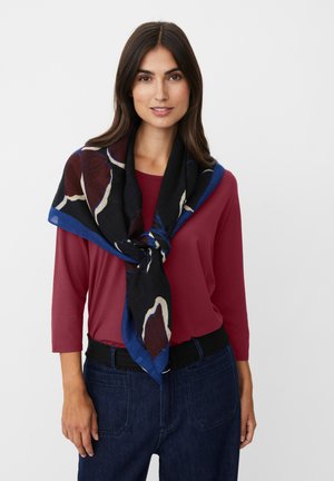 Frau trägt ein kastanienbraunes Oberteil, dunkelblaue Jeans und einen schwarzen Schal mit blauen und braunen Blättern, der um ihren Hals gebunden ist.