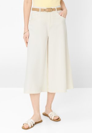 Frau trägt cremefarbene, weit geschnittene, verkürzte Hosen mit Seitentaschen, beigem Gürtel, gelbem Oberteil, goldenem Armband und weißen flachen Sandalen.