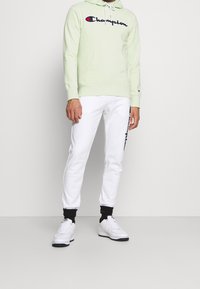 Ljusgrön hoodie med "Champion"-logotyp, matchad med vita sweatpants och strumpor med svarta detaljer. Vita sneakers fulländar outfiten.