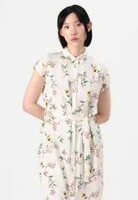 Blümchenhemdkleid aus cremfarbigem Stoff, mit kurzen Ärmeln, einem kragenförmigen Ausschnitt, Knopfverschluss vorne und einem passenden gebundenen Gürtel, verziert mit bunten Blumen.