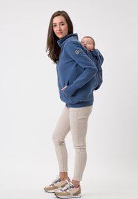 Felpa blu a conchiglia morbida per il portare i bambini, con colletto alto, tasche frontali e un supporto per il neonato sul retro, indossata con pantaloni beige chiaro e sneakers.