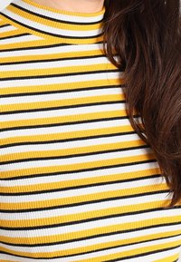 Top de canalé con rayas verticales en amarillo, blanco y negro. Cuello alto y diseño ajustado. La textura es suave y tiene una apariencia blanda.