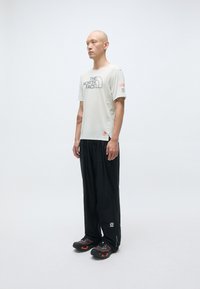 Chemise à manches courtes gris clair avec logo noir, associée à un pantalon noir ample. Chaussures de sport noires et orange.