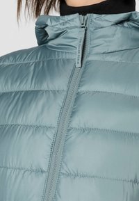 Světlá modrá péřová bunda s vysokým límcem a zipem. Vyznačuje se prošívanou strukturou s horizontálními švy. Hladký, lesklý materiál.
