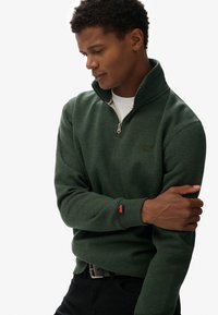 Groene kwart-rits sweatshirt met een gestructureerde afwerking, voorzien van een klein logo en een contrasterende rode accent op de manchetten. Zwarte jeans zichtbaar.