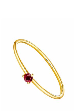 Anillo delgado de oro con una pequeña piedra preciosa roja redonda engastada en un engaste de cuatro garras sobre un fondo blanco.