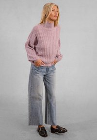 Donna in piedi con le mani nelle tasche, indossa un maglione girocollo a coste rosa, jeans a gamba larga sbiaditi e mocassini in pelle nera.