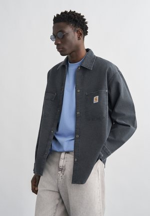 Mand iført runde solbriller, lyseblå skjorte, mørkegrå jakke med knapper og brystlommer samt lysegrå bukser, stående mod en hvid baggrund.