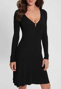 Robe noire côtelée à manches longues, décolleté en V, avec un détail froncé au niveau de la poitrine. Le tissu a une finition texturée et s'évase légèrement à l'ourlet.