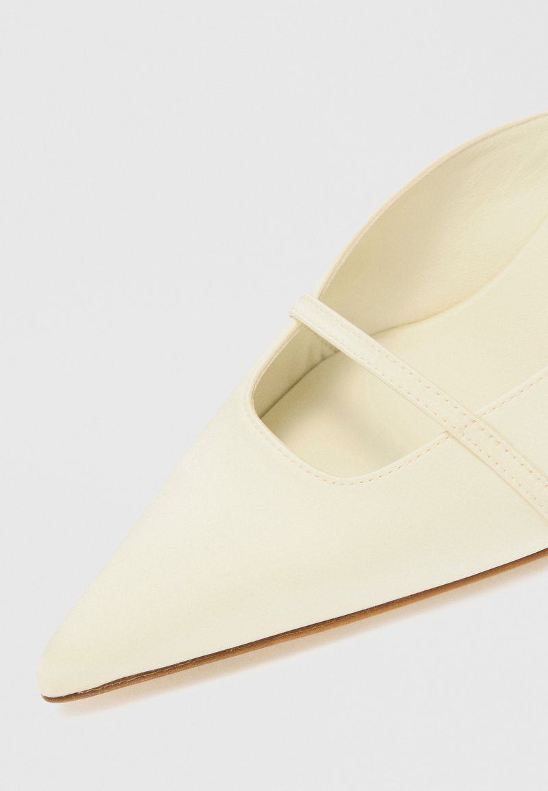 Zapato de punta en suave cuero crema, con un diseño de recortes y una correa delgada. La suela es de un marrón claro, mostrando una costura mínima.