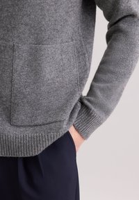 Szary wełniany sweter z prążkowanym mankietem i dołem, z dużą przednią kieszenią. Tekstura wydaje się miękka, z drobnym splotem dzianiny.