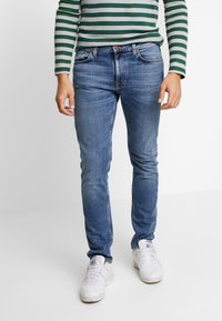 Jeans skinny de mezclilla azul con un acabado desgastado, cinco bolsillos y cierre de botón, combinados con una camisa de manga larga a rayas en verde y gris.