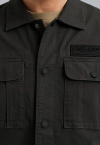 Veste utilitaire gris foncé avec deux poches avant, fermetures à boutons et un patch logo brodé. Fabriquée en tissu robuste avec une finition texturée.