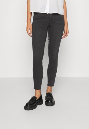 Personne portant un jean skinny noir, des mocassins noirs épais avec des embellissements, et une chemise blanche boutonnée, debout sur un sol gris.