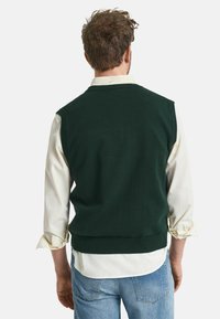 Donkergroene mouwloze trui gedragen over een witte lange mouw shirt, met een geribde zoom en een zachte textuur, gecombineerd met lichtblauwe jeans.