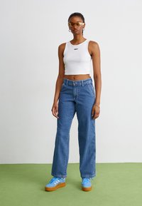 Baltas trumpas marškinėlių sluoksnis su "OBEY" logotipu, mėlyni plačiakojai džinsai ir mėlyni platforminiai sportbačiai su rudomis padais. Skaidrios geltonos saulės akys.