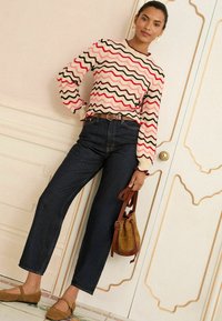 Das Model steht mit einer Hand auf der Hüfte, trägt einen pullover mit wellenförmigem Muster, dunkle Jeans und braune Ballerinas und hält eine braune Tasche. Eleganter Hintergrund.
