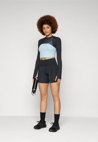 Black long-sleeve top over a light blue cropped top, paired with black shorts. The model holds a black water bottle, wearing black shoes and socks.