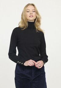 Pull à col roulé noir en matériau doux, avec des manches longues ornées de bouton doré aux poignets. Design en maille texturée.