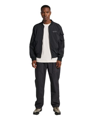 Mężczyzna stoi frontem, ubrany w czarną kurtkę na zamek z napisem „HALO”, białą koszulkę, czarne spodnie i beżowe buty trekkingowe na białym tle.