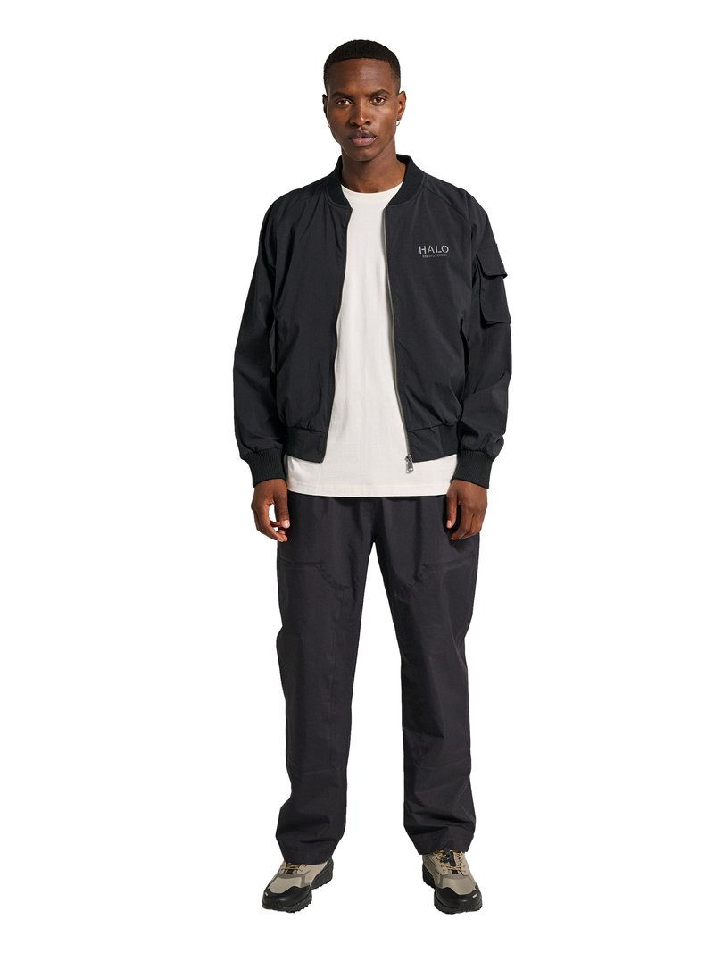 Mand står vendt fremad iført en sort lynlåstrøje med teksten "HALO", hvid T-shirt, sorte bukser og beige vandresko på hvid baggrund.