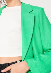 Blazer verde hecho de tela texturizada con solapas con muesca, combinado con un top corto blanco ajustado. Acento de un detalle floral en pantalones oscuros.