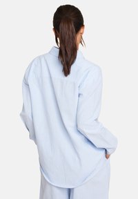 Lyseblå, langermet skjorte laget av myk stoff med kragehals, buet nederkant og avslappet passform. Har ryggstykke og sidelommer.