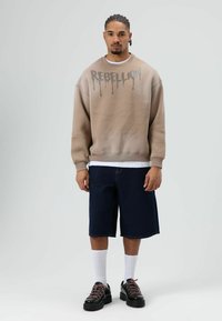 Beige sweatshirt met de tekst "REBELLION" in grijze druppelende letters, gecombineerd met donkere denim shorts. Model draagt witte sokken en zwarte chunky sneakers.