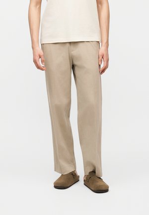 Mężczyzna w beżowych prostych spodniach, brązowych zamszowych butach wsuwanych i białej koszuli z krótkim rękawem, stojący na jednolitym tle.