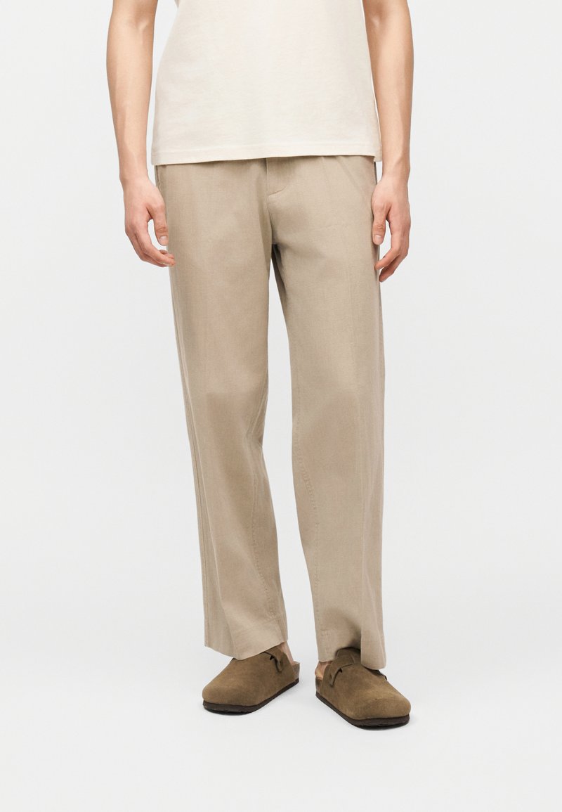 Άνδρας που φοράει μπεζ παντελόνι με ευθείες γραμμές, καφέ σουέντ παπούτσια χωρίς κορδόνια και λευκό κοντομάνικο πουκάμισο, στέκεται μπροστά σε απλό φόντο.