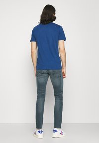 Man med axellångt mörkt hår som bär en blå t-shirt, slitna tajta jeans och vita sneakers med blå och röda detaljer, ståendes mot en vit vägg.