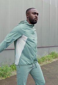 Helesinine spordijakk valgete aktsentidega, lukuga tasku ja kapuutsiga, koos sobivate pükstega, inimesel välitingimustes.