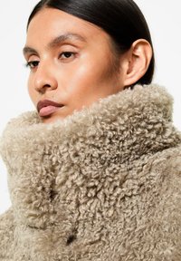 Donna con pelle liscia e capelli scuri che indossa un cappotto beige in finta shearling a collo alto e strutturato, che guarda di lato su uno sfondo bianco.