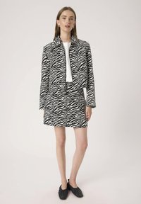 Frau, die ein schwarz-weiß gestreiftes Zebra-Muster-Jacket mit passendem Rock und einem weißen Top sowie schwarzen flachen Schuhen trägt, vor einem einfarbigen Hintergrund stehend.
