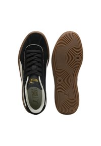 Zapatillas de ante negro con acentos blancos, suela de goma texturizada con patrones circulares y branding dorado. Se incluyen dos vistas: superior y de la suela.