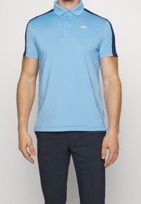 Ljusblå pikétröja med enfärgad design, kontrasterande marinblåa ärmar och en liten vit logga på bröstet. Kortärmad med spridd krage.