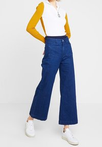 Wide-leg blue denim jeans with a high waist, front pockets, and a button closure, paired with a white and mustard long-sleeve top.