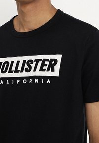 Černé bavlněné tričko s bílým vyšívaným obdélníkovým logem s nápisem "HOLLISTER" a "CALIFORNIA" v tučných písmenech. Krátké rukávy.