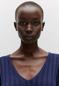 Dunkelhäutige Person mit kurzem Haar, die ein navyblaues gestreiftes V-Ausschnitt-Top trägt. Hat ausgeprägte Schlüsselbeine und einen neutralen Hintergrund.