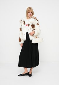 Femme aux cheveux blonds portant un manteau en fausse fourrure blanc et marron, un top blanc, une jupe plissée noire, des ballerines noires et tenant un petit sac à main blanc.