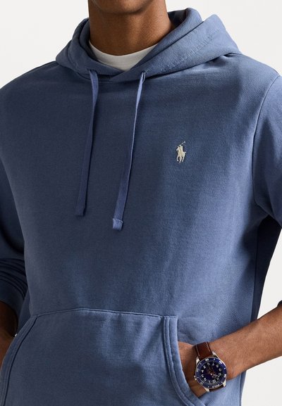 Blauwe katoenen hoodie met een voorzak, een capuchon met trekkoorden en een klein wit logo. Gecombineerd met een zilveren en bruine horloge met een blauwe wijzerplaat.