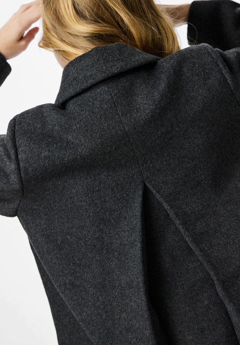 Kolgrå ullkappa med en bred krage, texturerad tyg och en avslappnad passform. Har sidofickor och en lätt draperad baksida.