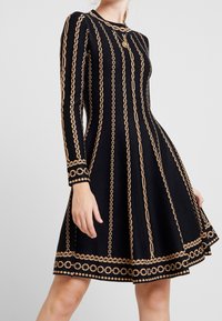 Abito a maglia nero con maniche lunghe, gonna a campana e dettagli a catena dorata lungo le cuciture verticali e i polsini.
