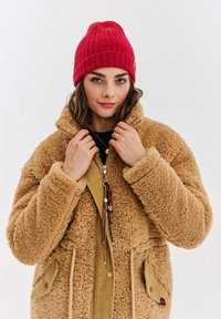 Žena v červené pletené čepici a béžovém teddy kabátu, držící límec kabátu proti jednoduchému bílému pozadí.