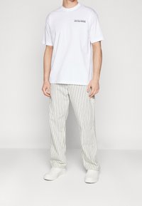 Mężczyzna w białej koszulce z małym napisem "Na wakacjach" oraz luźnych białych spodniach z cienkimi pionowymi czarnymi paskami, stojący z odprężonymi rękami.