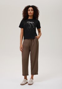 Sort t-shirt med korte ærmer og en sløjfe-design lavet af hvide udsmykninger, parret med løse, brune, ankellange bukser og beige sneakers.