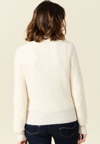 Kermanvärinen neule, jossa on pehmeä, ribbikuviointi ja pyöreä pääntie. Varustettu pitkillä hihoilla ja hieman lyhentyneellä helmalla.