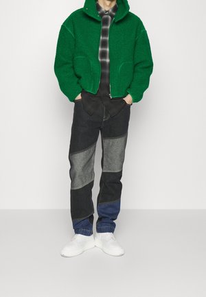 Personne portant une veste à capuche verte texturée sur une chemise à carreaux noir et blanc, un jean patchwork et des baskets blanches, les mains dans les poches.