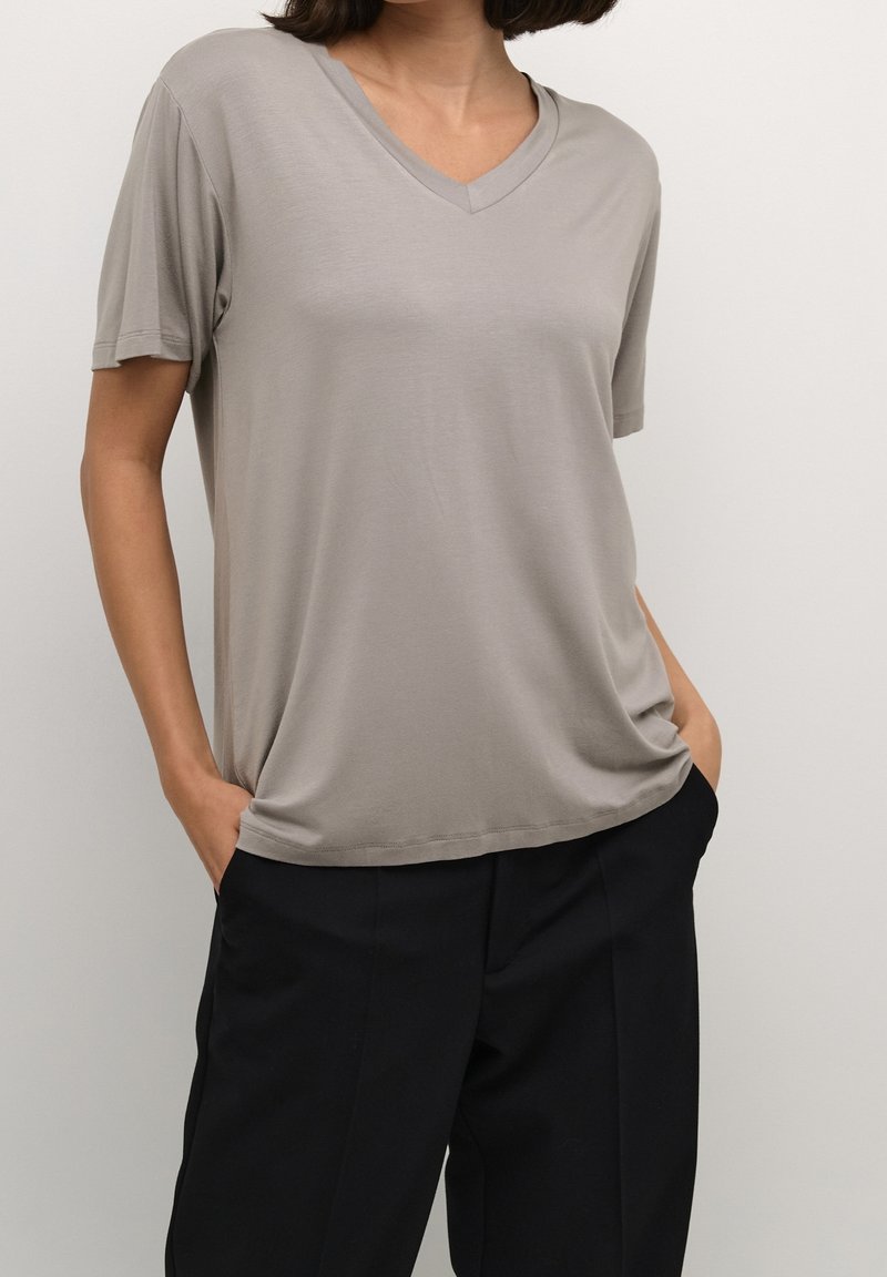 Femme portant un t-shirt gris clair à col en V et manches courtes ainsi qu'un pantalon noir, debout les mains dans les poches devant un fond uni.