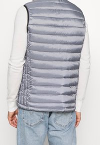 Graue Pufferweste mit horizontalen Nähten, hohem Kragen und ärmellos. Über einem weißen Pullover getragen, kombiniert mit hellblauen Jeans.